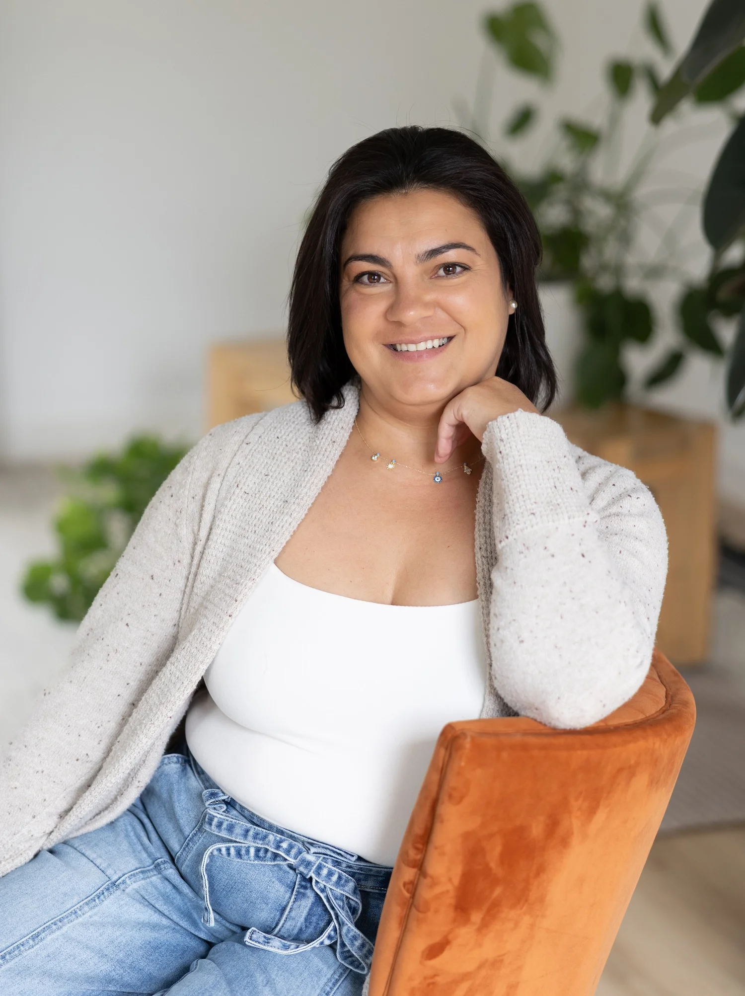 Dr. Tatiana Turo-Handy sitting in a wooden chair, smiling, with green plants and a light-colored room behind her.