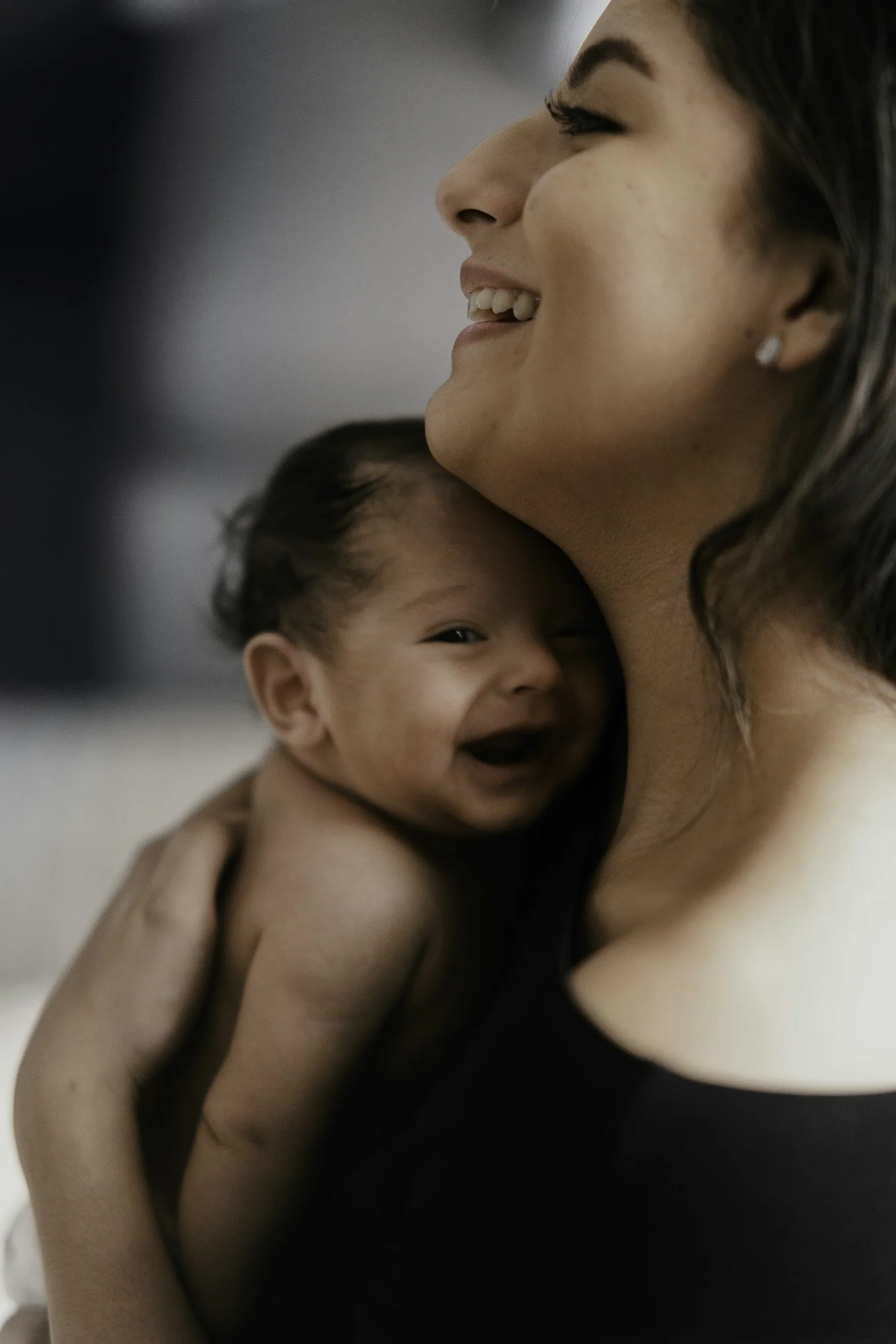 A woman smiling and hugging a toddler, with the toddler also smiling and showing a happy expression, together in an indoor setting.